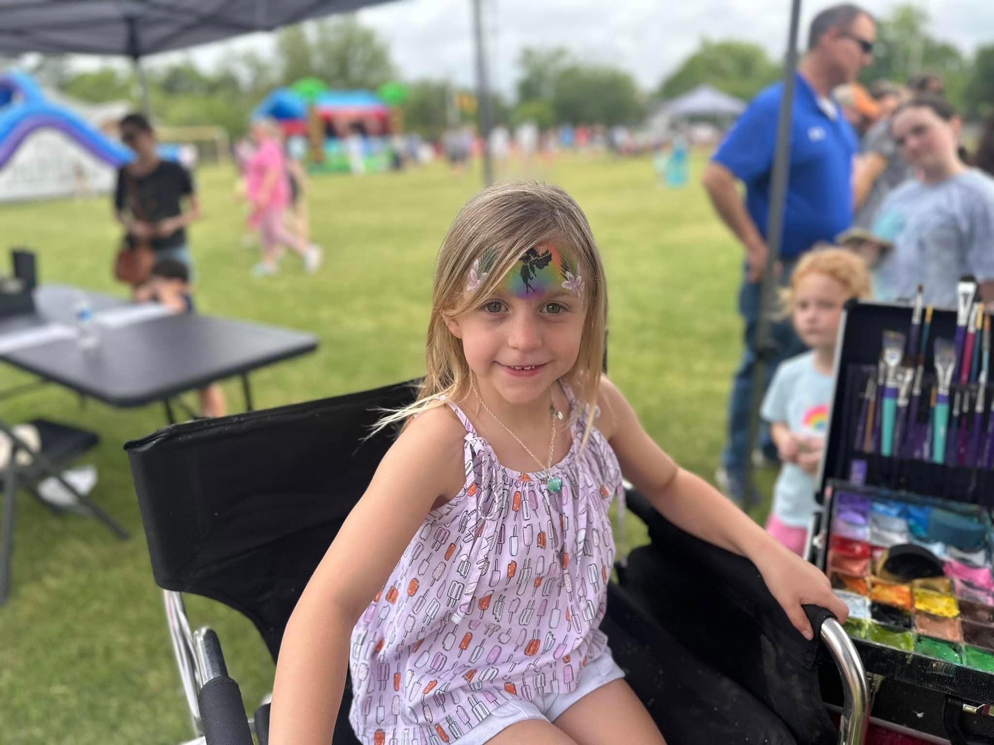A child smiling in a face painting chair at an outdoor event